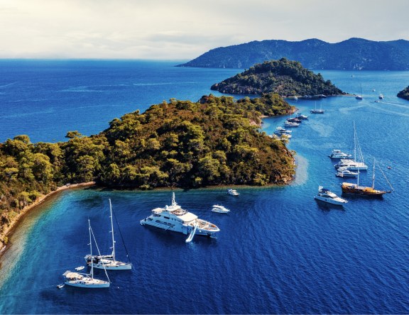 A breathtaking aerial view of a tranquil bay along the Turkish coastline, featuring lush green islands surrounded by crystal-clear turquoise waters. The bay is dotted with a variety of yachts and sailboats, anchoring near the shore or navigating the calm sea, illustrating a popular and picturesque yachting destination.