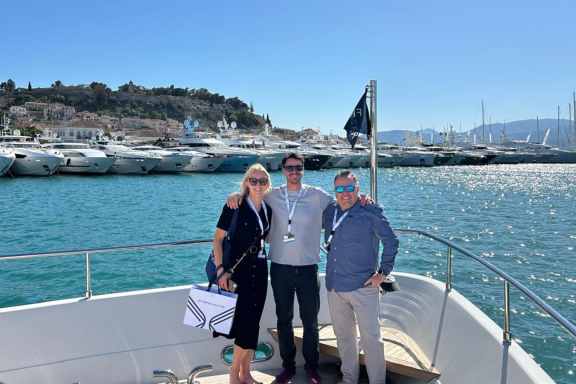 Charter Broker Sarah Klische, Louis Lamprell and Chris De Kock standing on the bow of a yacht with a stunning marina backdrop. On the left, a woman wearing a dark nautical-themed dress and sunglasses smiles alongside two men. The man in the middle, dressed casually in a light shirt and jeans, and the man on the right, in a blue shirt and sunglasses, both appear relaxed and happy. The marina behind them is filled with a variety of yachts under a clear blue sky, reflecting the sparkling waters, with a charming hillside town in the distance.
