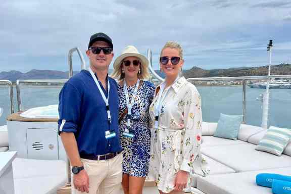 Charter Broker Daniel Asmus, Martina Asmus, and Nadja Asmus enjoy a sunny day aboard a yacht. The man on the left wears a navy blue cap and shirt complemented by beige trousers, while the middle woman wears a wide-brimmed hat and a patterned dress. The woman on the right sports sunglasses with a white floral dress. They all appear relaxed and cheerful, posing on the yacht's deck with a scenic marina and hilly landscape in the background.